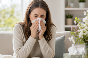 Junge Frau niest in ein Taschentuch in heller Wohnumgebung – Symbolbild für Allergie, Heuschnupfen und allergische Beschwerden.