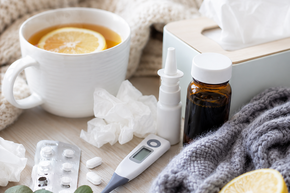 Tee mit Zitrone, Medikamente, Nasenspray und Thermometer auf einer Decke – Symbolbild für Grippe, Erkältung und häusliche Behandlung von Krankheitssymptomen.