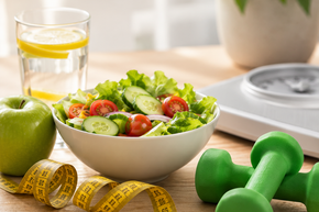 Salat, Maßband, Hanteln und Waage auf einem Tisch – Symbolbild für gesundes Abnehmen, Ernährung, Bewegung und Gewichtsreduktion.